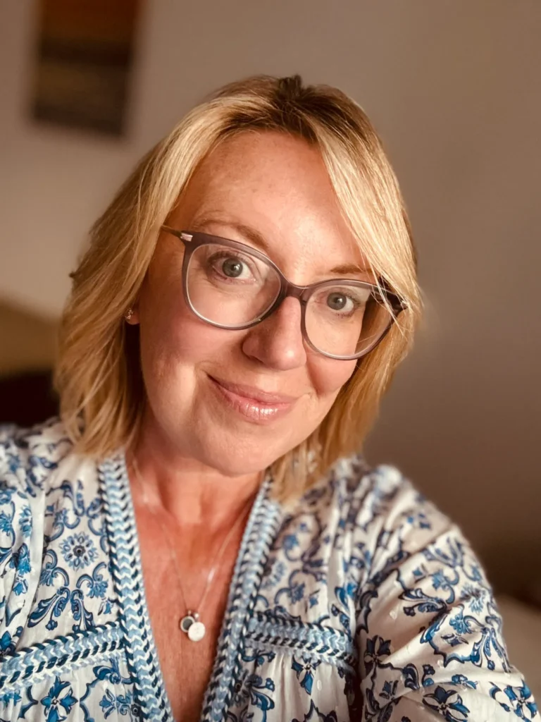 Helen, a white women with glasses and short wavy blonde hair, smiles at the camera. She is wearing a white and blue shirt. Helen helps Overwhelmed Business Owners claim back time