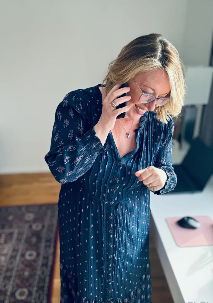 Helen, offering Executive Virtual Support, smiles while holding her phone to her ear stood at the desk. She has a long blue dress on and short wavy blonde hair.