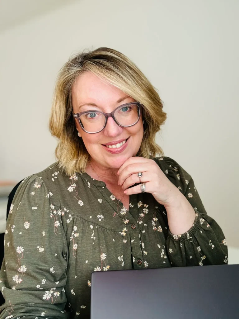 Professional virtual assistant Helen from Aurora Virtual Solutions, smiling warmly while seated at her desk. She is wearing glasses and an olive-green floral blouse, with a laptop in the foreground, representing tailored virtual assistant services for overwhelmed business owners.