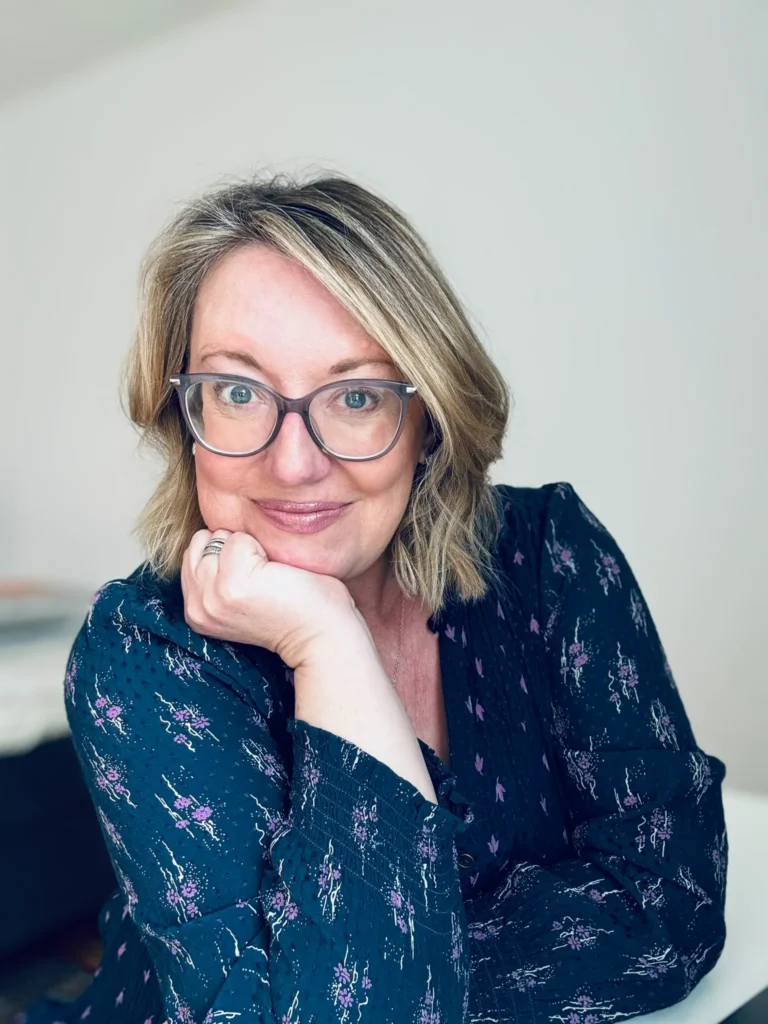 Helen, a professional virtual assistant from Aurora Virtual Solutions, smiling warmly while resting her chin on her hand. She is wearing glasses and a navy blouse with a delicate floral pattern, sitting in a bright and minimalistic workspace.